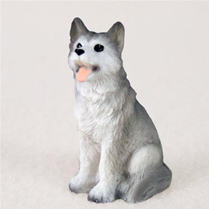 A small, detailed figurine of a gray and white Siberian husky dog sitting in an upright, loyal position. The miniature sculpture features a textured surface that realistically mimics thick fur, with dark gray coloring on its ears, back, and tail, while its face, neck, and underbelly are a clean white. The dog has a friendly and alert expression, with its mouth slightly open and a small pink tongue sticking out. Its dark brown eyes look upwards and to the left, adding a sense of personality to the piece. This figurine is designed as a decorative accessory for a bonsai tree, terrarium, or miniature garden, shown here against a plain, off-white background.