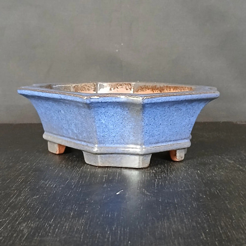 A 7-inch blue glazed Yixing bonsai pot, model No. 2411H, is centered in the frame against a muted gray background. The pot features a classic hexagonal shape with six equal sides that flare gracefully outward from the base to a wide, flat-topped rim. Its exterior is coated in a vibrant, mottled blue glaze with a fine, speckled texture, while silvery-gray highlights accentuate the sharp edges and corners of the vessel. The pot is elevated by four small, blocky feet that show the natural, unglazed reddish-brown Yixing clay on their bottom surfaces. The interior of the pot remains unglazed, revealing the same earthy terracotta-colored clay. The entire piece sits on a dark, textured surface that resembles a black wood grain, with soft light reflecting off the glossy glaze and the surface in front of the pot.