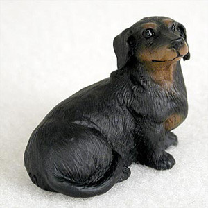 A detailed, miniature figurine of a black and tan dachshund is shown sitting patiently on a neutral, off-white surface. This small decorative accessory is designed to be placed at the base of a bonsai tree or within a terrarium, adding a touch of personality to indoor garden displays.

The figurine captures the characteristic features of a long-haired dachshund, with wavy, textured fur sculpted across its body. It is primarily black with classic tan markings on its muzzle, chest, and small "eyebrow" spots above its dark, glossy eyes. The dog is posed in a seated position, looking slightly upward and to the right with a calm and loyal expression. The realistic detailing of its paws and slightly curled tail makes it an endearing addition to any miniature landscape or bonsai collection.