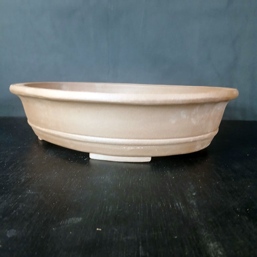 A side-view photograph of a 16-inch oval, unglazed Yixing bonsai pot, showcasing its minimalist and elegant design. The pot is crafted from traditional Yixing clay, featuring a warm, earthy tan or light brown color with a smooth, matte finish. It has a distinctive flared rim that curves slightly outward and a single horizontal decorative line encircling the lower half of the body. The pot is elevated by four small, integrated rectangular feet, which provide essential drainage and airflow for a bonsai tree. It is positioned on a dark, textured black wooden surface against a neutral, dark gray background, which makes the pot’s clean lines and natural clay color stand out. This professional-grade ceramic container is designed for aesthetic appeal and the health of medium-to-large bonsai specimens.