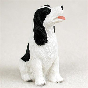 A small, detailed figurine of a black and white English Springer Spaniel, designed as a decorative accessory for a bonsai tree. The miniature dog is shown sitting upright, looking up with a cheerful expression, its mouth slightly open and a pink tongue visible. It features the breed's characteristic long, wavy fur, represented by a sculpted texture. The figurine is primarily white with prominent black markings on its head, long floppy ears, and back, creating a realistic and charming miniature scale model for a desktop bonsai or fairy garden.
