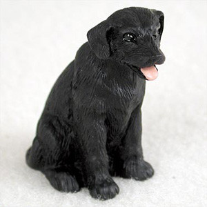 A detailed miniature figurine of a black Labrador Retriever puppy, designed as a decorative accessory for a bonsai tree or fairy garden. The figurine is shown in a seated position, with its head turned slightly to the right in an alert and friendly pose. It features a cheerful expression with its mouth open and a small, pink tongue sticking out. The sculpture has a realistic, textured surface that mimics the look of short fur, and its dark, glossy eyes catch the light. Set against a soft, out-of-focus white background, this charming black lab miniature captures the playful and loyal essence of the breed, making it a perfect addition to a miniature landscape or pet-themed display.