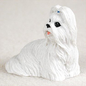 A detailed miniature white Shih Tzu figurine, designed as a decorative accessory for a bonsai tree or indoor garden from Bonsai Outlet. The figurine is depicted in a seated pose, facing toward the right, with its long, bright white fur meticulously textured to show flowing individual strands. Its characteristic Shih Tzu features include large, glossy black eyes, a small black nose, and a tiny pink tongue peeking out from its mouth. A small blue ribbon is tied at the top of its head, gathering its long facial hair into a classic topknot. The entire piece sits on a neutral, off-white textured background, emphasizing its clean white color and the intricate craftsmanship of the mold.