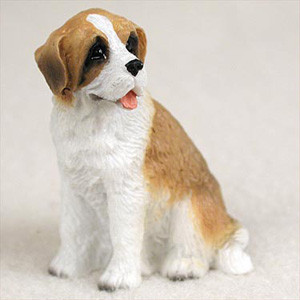 A detailed miniature figurine of a Saint Bernard puppy with a rough coat, designed as a decorative accessory for bonsai trees or miniature gardens. The small puppy is shown in a sitting posture, looking slightly to its right with an alert and friendly expression. It features realistic coloring with a white chest, paws, and muzzle, a white stripe running down its face, and warm tan-to-brown patches on its ears, around its eyes, and across its back. The figurine’s surface is finely textured to represent the thick, coarse fur of a rough-coated Saint Bernard. Its small pink tongue is visible in its slightly open mouth, complementing its dark, detailed eyes and black nose. This charming figurine is photographed against a neutral, light-colored background that emphasizes its intricate details and lifelike appearance.