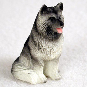 A detailed, hand-painted miniature figurine of a Keeshond dog, designed as a decorative accessory for a bonsai tree or miniature garden. The figurine is captured in a classic sitting pose with an alert, friendly expression and a small pink tongue slightly visible as if panting. 

The figurine accurately depicts the breed's thick, double coat with molded fur textures. The coloring is a gradient of charcoal and silver-grey along the ears, back, and shoulders, fading into a clean, creamy white on the ruff, chest, and legs. It features the breed's distinctive dark markings around the eyes, often called "spectacles." This small Keeshond figurine is intended to add a sense of life, scale, and personality when placed at the base of a bonsai tree. The figurine is shown resting on a soft, textured white surface.