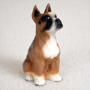 A detailed miniature figurine of a fawn Boxer dog, designed as a decorative accessory for a bonsai tree landscape. The small dog is captured in an upright sitting pose with its chest out and its head tilted up, looking alertly toward the right. Its rich brown coat is accented with crisp white markings on its paws, broad chest, and muzzle, and it features a distinctive black mask around its dark eyes and nose. The figurine's pointed ears stand tall, giving it an attentive and loyal expression. The piece is shown resting on a soft, light-colored textured surface against a neutral, out-of-focus background, highlighting the craftsmanship of the miniature.