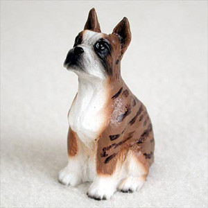 A detailed miniature figurine of a brindle Boxer dog, designed as a decorative accessory for a bonsai tree landscape. The small statue depicts the dog in an attentive sitting pose, with its head tilted slightly upward and to the side. It features realistic breed details such as a square muzzle, alert upright ears, and a classic brindle coat pattern with dark black stripes over a tan-brown body. The figurine also has a bright white chest, white front paws, and a white blaze on its face. The craftsmanship highlights a subtle textured surface to mimic short fur, making it a lifelike and charming addition to a miniature garden, terrarium, or indoor bonsai display.