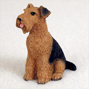 A detailed miniature figurine of an Airedale Terrier dog, designed as a decorative accessory for bonsai trees. The dog is depicted in a sitting position, looking up and to the left with its small pink tongue slightly visible. It features the breed's characteristic wiry-textured coat, with a warm tan color covering most of its body and a prominent black saddle mark on its back and tail. The figurine is realistically sculpted to show individual tufts of fur and sits against a neutral, plain off-white background.