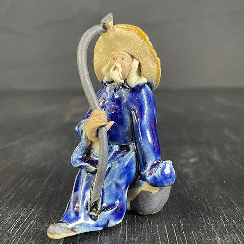 A small, glazed ceramic Chinese "mud man" figurine designed as a decorative accent for bonsai trees or miniature gardens. The figure depicts an elderly man with a serene expression, a long white beard, and a mustache. He is dressed in a flowing, deep blue glazed robe with a high-shine finish that shows variations in color where the glaze has pooled. 

On his head sits a wide-brimmed, textured hat in a matte yellowish-tan color. The man is in a seated position, holding a long, slender, and curved grey staff with both hands. The figurine is photographed from a slight side angle, resting on a dark, weathered wood surface against a neutral dark grey background, highlighting its handcrafted details and vibrant colors.