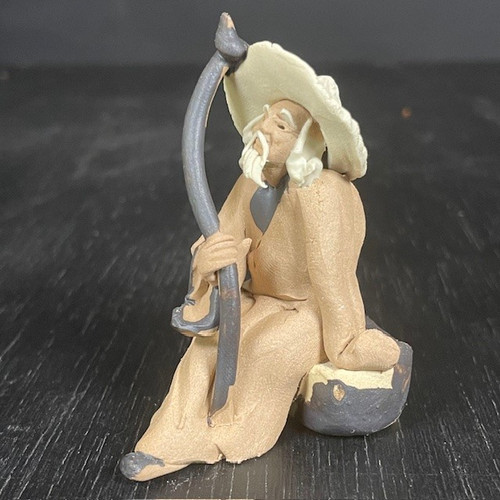 A detailed photograph of a traditional unglazed Chinese clay figurine, often referred to as a "mudman," depicting an elderly man sitting in a peaceful pose. The figure is dressed in a long, tan-colored robe and wears a large, wide-brimmed, cream-colored hat. He has long, flowing white facial hair, including a prominent mustache and beard that rest against his chest. In his right hand, he holds a tall, slender, dark grey staff that curves gracefully above his head. The man is perched on a small, dark grey, rock-like base. The entire sculpture has a rustic, handcrafted appearance with a matte, unglazed texture characteristic of classic bonsai figurines. This decorative miniature is designed to be placed within a bonsai tree arrangement to add a sense of scale, wisdom, and traditional storytelling. The figurine is photographed on a dark, textured wooden surface against a dark, blurred background, highlighting its neutral tones and fine details.