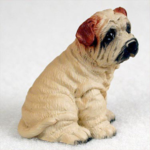 A detailed miniature figurine of a cream-colored Shar-Pei puppy, designed as a decorative accessory for a bonsai tree arrangement. The puppy is captured in a sitting position, facing slightly toward the right. The figurine prominently features the breed's signature deep skin folds and heavy wrinkles covering its entire body, from its neck down to its paws. The primary color of the figurine is a soft cream beige, accented by a dark black muzzle and black shading around its small, dark eyes. Its characteristic small, folded ears are painted in a contrasting reddish-brown tone. The puppy's short, thick tail is curled slightly at the base of its back, and tiny black nails are visible on its front paws. The entire figurine is set against a plain, off-white, textured background, highlighting its hand-painted details.