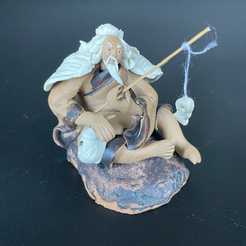 A detailed clay bonsai figurine of an elderly Chinese fisherman, often called a mudman, sitting on a textured dark rock. The figure has long, flowing white hair and a prominent white beard. He is dressed in a traditional tan-colored robe with dark brown trim and a white shawl draped over his shoulders. He holds a thin wooden fishing rod in one hand, with a white string and a small white clay fish dangling from the tip. His other hand rests on a larger white fish near his lap. The figurine is handcrafted with a matte, earthy finish and is set against a solid dark background, highlighting its intricate details for use as a bonsai decoration.