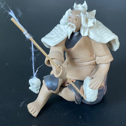 A detailed, handcrafted Chinese ceramic figurine of an elderly man, often referred to as a 'mud man' used for bonsai decoration. The figurine portrays a man sitting in a hunched, contemplative pose, engaged in the act of fishing. He holds a thin, light-colored wooden rod with a delicate white thread hanging from its tip, ending in a small white sculpted piece representing a catch or bait.

The man's features include small, concentrated eyes, a long pointed white beard, and a mustache. His head is crowned with a thick, textured mane of white hair that flares out like a stylized cloud. He is dressed in traditional attire consisting of tan-colored robes, a white cape-like garment with a ridged texture draped over his shoulders, and dark indigo-colored pants. His bare feet are visible as he sits. The sculpture is set against a solid dark grey background, highlighting its earthy tones and intricate handcrafted textures.