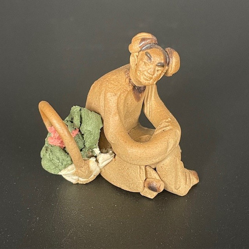 A detailed ceramic figurine of a Chinese woman sitting on the ground, crafted from unglazed, reddish-brown clay with a slightly textured, matte finish. The woman has a joyful, smiling face with eyes narrowed and dark painted eyebrows, and her hair is styled in two small buns on her head. She is shown in a relaxed, seated pose with one arm resting on her lap, while her other arm reaches toward a small basket at her side. The basket is filled with clumpy, textured clay elements in green and pink, representing a fresh bouquet of flowers. This miniature figurine, often used as a bonsai or terrarium accessory, captures a moment of traditional rural life. The piece is set against a solid dark grey background, highlighting the earthy tones and handmade details of the oriental mudman-style art.