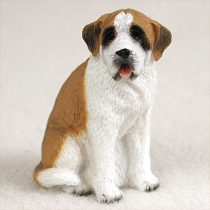 A detailed miniature figurine of a Saint Bernard with a smooth coat, perfectly sized for placement within a bonsai tree landscape. The dog is depicted in a seated position, looking forward with a friendly expression and its mouth slightly open, revealing a tiny pink tongue. Its coat features classic breed markings: a predominantly white body and chest, a large tan-brown patch across its back, and tan ears. Its face has a prominent white blaze down the center, framed by black markings around its dark eyes and a black nose. The molded texture of the figurine gives the appearance of short, dense fur, adding to its realistic charm against a neutral, off-white background.