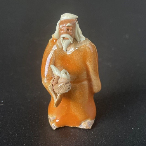 A detailed studio shot of a traditional Chinese mudman figurine, a popular ceramic accessory for bonsai trees. This small, handmade clay figure depicts an elderly standing man with a contemplative expression. His face and hands are made of unglazed reddish-brown clay, featuring a long, flowing white beard and mustache that match his white hair and cap. He wears a brilliant, glossy orange-glazed robe that reflects the light. In his hands, he holds a long, pale-colored pipe, caught in the act of smoking. The figurine is positioned against a solid dark gray background, emphasizing the rich colors and textures of the glazed and unglazed ceramic craftsmanship.