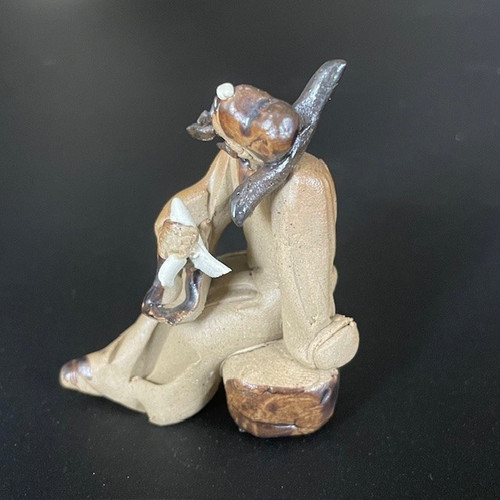 A small, handcrafted Chinese ceramic mudman figurine designed as a decorative bonsai accessory. The figurine depicts a man in a hunched, sitting position atop a dark brown, textured base that resembles a rock or tree stump. Crafted from light tan, unglazed clay, the figure features dark brown glazed accents on his hair and a long, dark object slung over his shoulder. He is leaning forward, holding a delicate, white-colored piece of clay in his hands, which represents a scarf. The artisanal, impressionistic style of this miniature sculpture adds a traditional feel and a sense of scale and narrative to a bonsai tree arrangement. The figurine is photographed from a side-profile view against a neutral, dark gray background.
