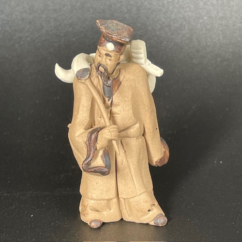 A detailed miniature Chinese figurine of an elderly standing man, crafted from unglazed tan clay in a traditional "mudman" style. The sculpture depicts a man with a contemplative expression, characterized by a long, dark pointed beard and a drooping mustache. He is dressed in a simple, flowing tan-colored robe with subtle folds.

On his head, he wears a tall, dark brown hat featuring a single white circular emblem at the center of the brim. He is shown in a slightly hunched pose, leaning on a simple wooden staff held in his right hand. Over his back and shoulders, he carries a large, white, abstractly shaped object, possibly representing a sack of scrolls or a bundle of goods. His small feet, clad in dark shoes, emerge from beneath the hem of his long robe.

The unglazed ceramic material gives the figurine a matte, earthy texture, with the tan clay providing a natural look that contrasts with the dark brown highlights on the hat, facial hair, and shoes. This small decorative figure, set against a dark, textured background, is a classic bonsai accessory designed to add a sense of scale, history, and peaceful storytelling to a miniature garden or indoor plant display.