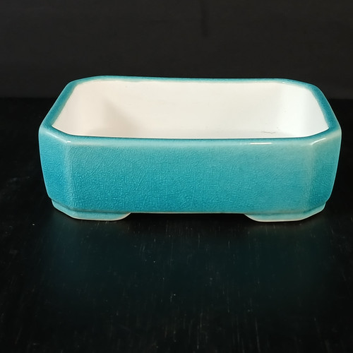 An eye-level, studio-lit photograph showcases a 5-inch glazed Yixing bonsai or succulent pot. The rectangular pot features a vibrant turquoise-blue exterior with a glossy finish and a fine, intricate crackle or crazing pattern throughout the glaze. The pot’s interior is a clean, contrasting matte white, and it is designed with softly rounded corners and small, integrated corner feet to provide drainage for plants. The container is centered on a dark, reflective black surface against a solid black background, which makes the bright teal color pop. This elegant ceramic piece is specifically designed for small bonsai trees or succulent arrangements.