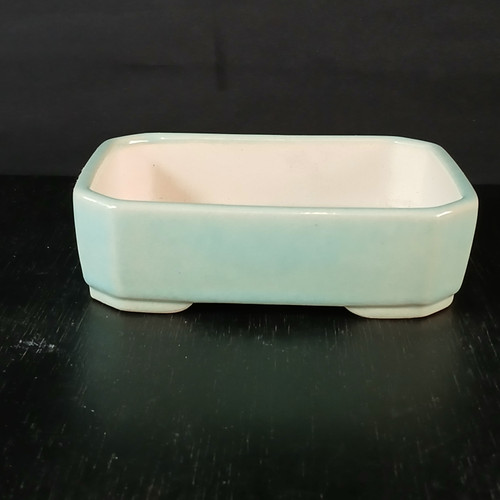 A 5-inch glazed Yixing bonsai or succulent pot sits centered on a dark, textured black surface against a solid black background. The rectangular pot features a smooth, glossy, pale mint green or celadon glaze on its exterior walls, which have softly rounded corners and a slightly beveled top rim. The interior of the pot is a contrasting pale cream color. It is supported by a raised, footed base in a matching off-white tone, with four distinct feet visible at the corners. The minimalist setting and high-contrast background emphasize the pot's simple, elegant design and delicate color.