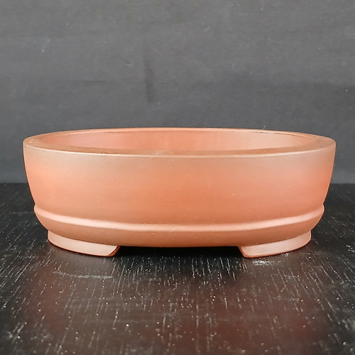A 4.5-inch, quality unglazed oval Yixing bonsai pot, showcased from a front-facing perspective. Crafted from fine, reddish-brown terracotta clay, the pot features a smooth matte finish and a classic oval shape with gently rounded sides. A subtle, elegant horizontal ridge wraps around the lower portion of the pot, just above its low-profile, integrated feet. The pot is positioned on a dark, textured wood-like surface against a solid dark gray background, emphasizing its clean lines and earthy tones. This Yixing pot (model YX1140) is a timeless and functional choice for displaying various small bonsai tree styles.