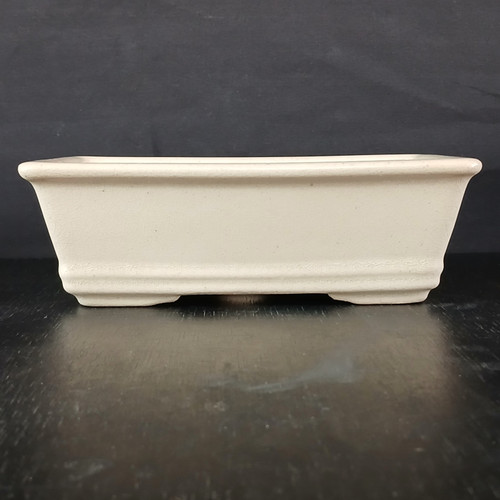 A side profile view of a 7-inch glazed cream rectangle Yixing ceramic bonsai pot. The rectangular pot has a clean, minimalist design with straight sides that taper slightly towards the bottom. It features a wide rim at the top and a decorative horizontal band near the base, which sits on four small, integrated corner feet. The creamy off-white glaze has a subtle crackle texture, giving the ceramic a vintage feel. The pot is empty and positioned in the center of the frame against a stark, solid black background, resting on a dark, reflective surface that highlights its simple and elegant form. This professional-grade Yixing pot is ideal for showcasing a variety of bonsai tree styles.