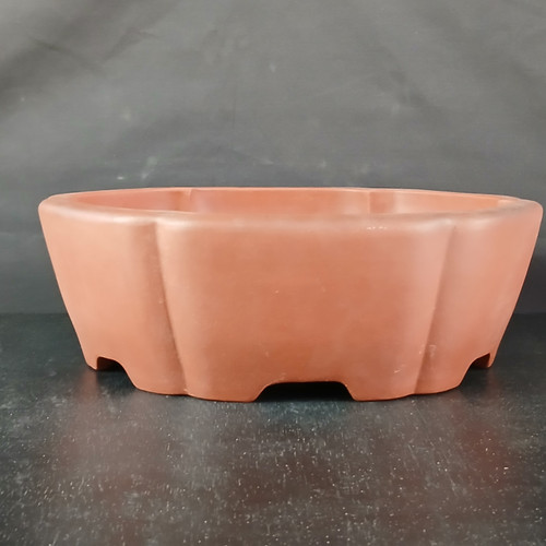 A front-facing eye-level photograph of a 13-inch round Yixing bonsai pot, listed as model No. 1784. The pot is crafted from unglazed, reddish-brown clay with a smooth, matte finish that highlights its natural, earthy texture. Its unique shape features elegantly scalloped or lobed sides, creating subtle vertical indentations that give it a "mokkoh" or melon-cross-section appearance. The pot has a flat, thick top rim and sits on small, integrated feet formed by shallow arches cut into the base, which allow for essential drainage and airflow for a bonsai tree. It is positioned on a dark, subtly speckled surface against a plain, dark gray textured background, with soft lighting that emphasizes its rounded curves and professional craftsmanship.