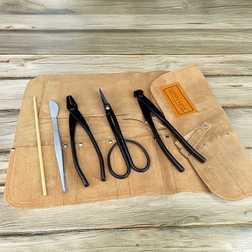 An overhead shot shows a Tinyroots professional-grade 5-piece bonsai tool set neatly organized within a light tan suede tool roll on a rustic, light-colored wooden surface. 

From left to right, the essential bonsai tools include a slender bamboo root pick for soil aeration; a stainless steel tool featuring fine-point tweezers on one end and a rounded spatula on the other; a pair of black carbon steel jin pliers for working with wire; long-handled black carbon steel bud shears for precision pruning; and a pair of black carbon steel concave cutters designed to create clean, flush cuts on branches. 

The high-quality suede tool roll features individual stitched pockets for each tool and a folding protective flap embossed with a rectangular leather "tinyroots" logo patch. This comprehensive set offers the foundational tools required for professional bonsai styling and maintenance.