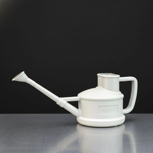 A side-profile studio photograph of a white Tinyroots watering can, specifically designed for the precise care of bonsai trees. The watering can features a minimalist, modern design with a long, slender spout that extends upwards at an angle, ending in a flared, rose-style nozzle designed for a gentle, controlled water flow. The main body consists of a compact, cylindrical reservoir with a raised, open-top filling port and a squared-off, D-shaped handle at the back. On the side of the body, a small rectangular plaque displays the embossed "Tinyroots" brand name. The object is set against a solid, dark gray-to-black background and rests on a smooth, metallic gray surface that shows a faint reflection of the can, highlighting its clean lines and functional aesthetic.