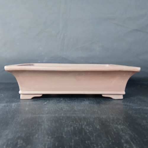 A side-view studio photograph of a 13.5-inch rectangular bonsai pot. The pot is crafted from unglazed Yixing clay, featuring a natural, matte, light brown or terracotta-colored finish. Its design includes elegant, outward-curving sides that lead to a wider top opening. The base is accented with a thin decorative ridge and rests on low, integrated rectangular feet with simple arched cutouts, providing both stability and space for drainage. The pot is displayed on a dark, slightly reflective surface against a neutral dark gray backdrop, highlighting its clean lines and earthy texture, which are well-suited for traditional bonsai aesthetics.