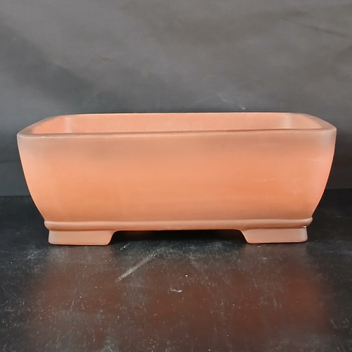 A front-facing, eye-level photograph of a 14-inch rectangular unglazed Yixing bonsai pot (model YX1094) from Bonsai Outlet. The pot is expertly crafted from authentic reddish-brown Yixing clay, exhibiting a smooth, matte, and non-reflective finish. Its classic rectangular design features soft, rounded corners and sides that subtly taper downward towards a decorative raised band just above the base. The vessel is supported by four solid, rectangular block-style feet at each corner, providing the necessary elevation for proper drainage and airflow essential for bonsai health. The pot sits on a dark, textured surface against a minimalist dark grey backdrop, which highlights its clean lines, warm earthy tones, and elegant proportions.