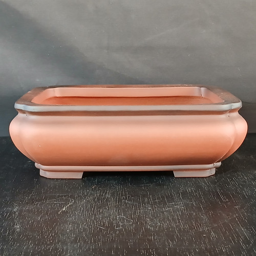 A 10-inch rectangular unglazed Yixing ceramic bonsai pot sits on a dark, weathered wooden surface. The pot is crafted from smooth, reddish-brown terracotta clay, featuring an elegant, bulbous profile with rounded corners and slightly convex sides. Its upper rim is finished with a subtle dark-charcoal smoke effect, creating a sophisticated contrast with the warm earthy tones of the main body. The pot is supported by four discreet, integrated corner feet, and its clean, recessed interior is visible. This traditional Yixing pottery piece is photographed against a dark grey backdrop, which highlights its classic lines and matte finish, making it a perfect choice for a mature bonsai tree.