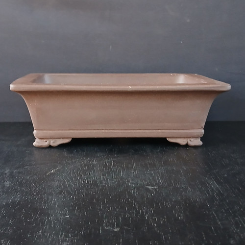 An eye-level studio photograph features a 10-inch unglazed rectangular Yixing ceramic bonsai pot, model No. 860. The pot is crafted from a traditional, warm reddish-brown clay with a smooth, matte finish that is characteristic of high-quality Yixing pottery. 

Its design showcases a prominent, outward-flaring rim at the top, which transitions into slightly inward-sloping walls down to a recessed base. The entire structure is elevated by four sturdy, decoratively carved feet that feature a subtle, cloud-like scroll pattern, adding an element of elegance and traditional craftsmanship. The pot is positioned on a dark, textured black surface that resembles aged wood, set against a seamless, dark gray background. This minimalist setting emphasizes the pot’s clean architectural lines and earthy tones, highlighting its suitability as a refined container for a mature bonsai tree.