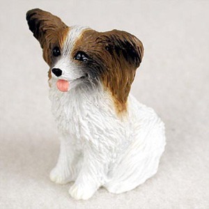 A detailed miniature figurine of a brown and white Papillon dog, designed as a decorative accessory for a bonsai tree or miniature garden. The small statue captures the breed's signature large, butterfly-wing ears in a rich brown color, which contrasts with the crisp white fur on its body and the white blaze running down its face. The Papillon is depicted in a sitting position with a happy expression, featuring bright black eyes, a small black nose, and a tiny pink tongue peeking out. The figurine has a realistic, textured finish to mimic the appearance of soft fur, making it a charming addition for collectors and indoor bonsai enthusiasts looking to add personality to their displays.