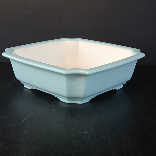 A professional studio photograph of a 9-inch light blue glazed square Yixing ceramic bonsai pot. The pot features a pale, serene celadon-like blue glaze with a glossy finish that catches the light. Its shape is square but with decorative indented corners, giving it an elegant, eight-sided profile. The rim is slightly flared outwards, and the pot is supported by four low, integrated feet at each corner. The unglazed interior shows a light, creamy ceramic body, characteristic of high-quality Yixing clay. Set against a stark black background on a dark, subtly reflective surface, the pot’s clean lines and soft color are emphasized, showcasing it as a premium choice for displaying a bonsai tree.