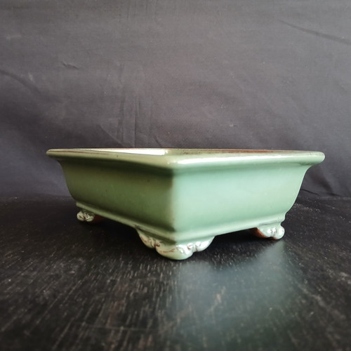A side-angle photograph shows a 7-inch rectangular glazed Yixing bonsai pot in a delicate pale green celadon color. The pot features a wide, lipped rim with a subtle brown edge where the glaze thins, and it is elevated by four ornate, cream-colored cloud feet. The glossy ceramic finish reflects soft light, showcasing the pot's clean lines and gentle curves. It rests on a dark, textured wooden surface against a dark gray, subtly wrinkled fabric background, which emphasizes the pot's soft color and elegant design.