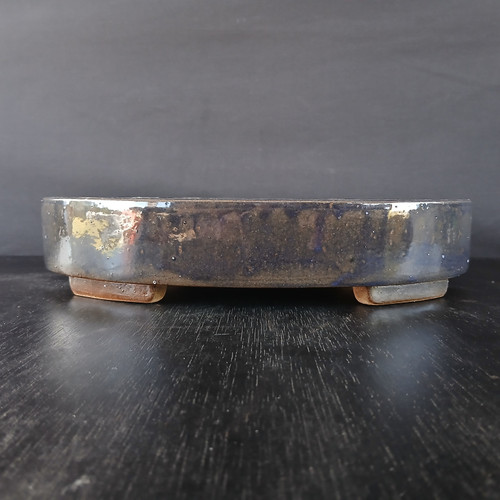 A profile view of a 12-inch glazed oval Yixing bonsai pot resting on a dark, wood-grain surface against a plain, dark gray background. The pot features a rich, dark glaze with a metallic, iridescent quality that reflects tones of deep blue, charcoal, and mottled patches of gold and bronze. Its low-profile, oval form has smooth, rounded edges and a level rim. The vessel is elevated on four small, unglazed rectangular feet in a light tan clay, which provides a natural contrast to the dark, glossy body of the ceramic. The lighting catches the reflective surface of the pot, highlighting its handcrafted texture and the depth of its glaze.