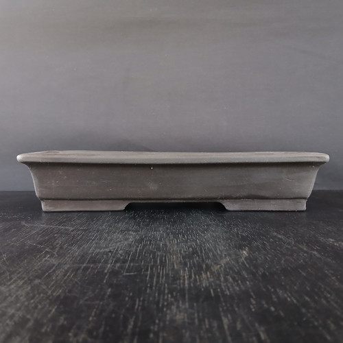 A horizontal, eye-level photograph shows a shallow, rectangular bonsai pot made from unglazed Yixing clay. The pot features a deep charcoal gray or brownish-black hue with a smooth, matte finish that highlights its clean lines. Its design includes a slightly flared rim and sides that taper inward toward the base. Along the bottom edge, thick, integrated feet elevate the pot, creating a wide, shallow arch underneath to facilitate drainage and air circulation for a bonsai tree's roots. The pot is centered in the frame, sitting on a dark, textured wooden surface with visible grain and wear, set against a plain, dark gray background that provides a neutral and focused setting.