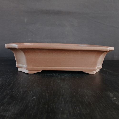 A detailed side-profile view of a premium 13.5-inch rectangular Yixing bonsai pot (model YX870A). Handcrafted from traditional reddish-brown unglazed ceramic, this high-quality pot features an elegant design with subtly notched corners and a flared top rim. A decorative horizontal ridge accents the lower portion of the pot, which is supported by four integrated, low-profile feet. The pot's matte, earthy texture and rich clay color are showcased against a minimalist dark grey backdrop, resting on a dark, reflective surface. The classic rectangular shape and breathable Yixing clay material make it a sophisticated choice for formal or informal upright bonsai tree styles.