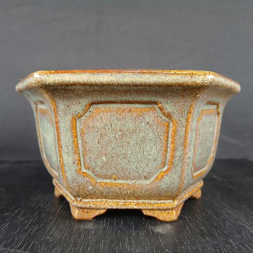 A studio photograph of a six-sided, hexagonal Yixing glazed bonsai pot from Bonsai Outlet. The pot is finished in a rustic, matte reactive glaze featuring a speckled dusty-blue and sage green body with warm, orange-brown highlights accentuating the edges and recessed areas. Each of the six side panels contains a decorative, octagonal-shaped inset frame with notched corners. The pot has a sturdy, slightly flared top rim and sits on six short, integral feet that follow the hexagonal shape of the base. This 6-inch pot is displayed on a dark wooden surface against a neutral dark grey background, emphasizing its earthy tones and handcrafted texture.