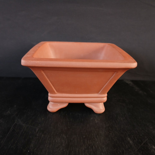 A 7-inch unglazed square cascade Yixing bonsai pot crafted from reddish-brown terracotta clay. The pot features a wide, flat rim and sides that taper downward toward a smaller base, creating the deep vessel characteristic of a cascade-style pot. Each of the four sides is decorated with a recessed panel and beveled edges, adding geometric depth. Near the bottom, two horizontal raised bands wrap around the base, which sits on four decorative, outward-curving feet. The pot is centered on a dark, textured black surface against a solid dark background, with soft lighting highlighting its matte finish and architectural details.