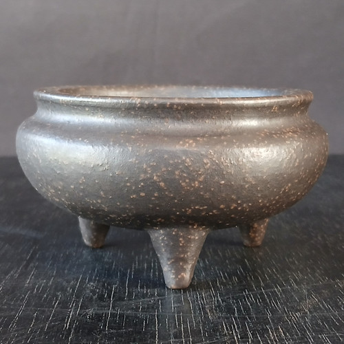 A close-up photograph of a small, unglazed round ceramic bonsai pot, model #1129-1. The pot features a bulbous, spherical body with a dark, charcoal-brown color and a matte finish. The surface is heavily textured with fine, light tan and golden-brown speckles that give it an earthy, stone-like appearance. It sits on three short, tapered tripod legs that support the wide, rounded base. A subtle, indented neck leads to a slightly flared rim at the top opening. The pot is centered on a dark, weathered wooden surface with visible grain patterns, set against a solid dark gray background. The soft lighting from the side highlights the organic curves and the gritty, unglazed texture of the clay, emphasizing its traditional and handcrafted aesthetic.