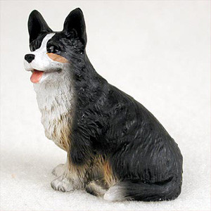 A detailed miniature figurine of a Cardigan Welsh Corgi dog, designed as a decorative accessory for a bonsai tree or indoor garden. The small dog is depicted sitting upright on its haunches, looking slightly to its left with an alert and happy expression. It features a predominantly black coat with a crisp white ruff around its neck and chest, a white blaze on its muzzle, and small tan accents near its cheeks and on its paws. Its large, pointed upright ears are characteristic of the breed, and its mouth is slightly open, showing a pink tongue as if panting happily. The figurine has a realistic, textured surface that mimics the appearance of thick fur, making it a charming and lifelike addition to any miniature bonsai landscape.