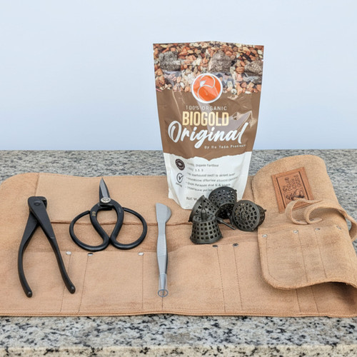 A flat-lay photograph showcases a professional bonsai care kit neatly arranged on a speckled gray and white granite countertop against a plain light background. 

In the center of the leather roll are three essential carbon steel bonsai tools: a pair of black concave cutters, large-handled black pruning shears, and a silver double-ended spatula and tweezer tool. These tools are resting on an open, tan leather tool roll that features multiple pockets and a fold-over flap with a branded leather patch. 

To the right of the tools are several small, black plastic mesh fertilizer baskets used for placing slow-release pellets on the soil surface. Standing behind the tools is a large brown and white bag of Biogold Original 100% organic fertilizer, showing its pellet-like contents in the top graphic. The arrangement suggests a comprehensive set of high-quality tools and supplies for maintaining and nourishing bonsai trees.