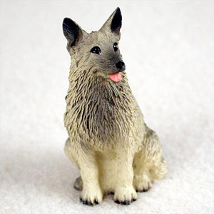 A small, detailed figurine of a Norwegian Elkhound dog, designed as a decorative bonsai tree accessory. The miniature statue is captured in a sitting position, looking alertly toward the side. It features a realistic, textured coat rendered in variegated shades of silver-grey and cream, with darker charcoal markings on the face and the tips of its pointed ears. The dog's expression is friendly, with dark, shining eyes and a small pink tongue peeking out from its mouth. This charming figurine is intended to be placed within a bonsai pot or miniature garden landscape, adding a sense of scale, life, and character to the Zen-like botanical display.