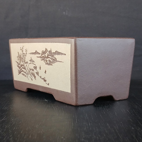 A 7-inch rectangular Yixing ceramic bonsai pot is shown from a three-quarter angle, resting on a dark wooden surface against a dark gray background. The pot is made from a high-quality, unglazed dark brown clay with a fine, matte texture. 

The focal point of the pot is a light cream-colored rectangular panel on the front side, featuring an intricately etched traditional landscape. This scene depicts a serene vista with distant mountains, a small pavilion nestled among stylized trees, and what appears to be water with small boats or reflections, all rendered in a dark brown ink-like style. 

The pot's design is clean and architectural, with straight sides and small, arched cut-outs at the bottom that form minimalist feet, providing a slight elevation. This elegant and traditional container is designed to complement the artistic form of a bonsai tree, offering a classic aesthetic suitable for various species.