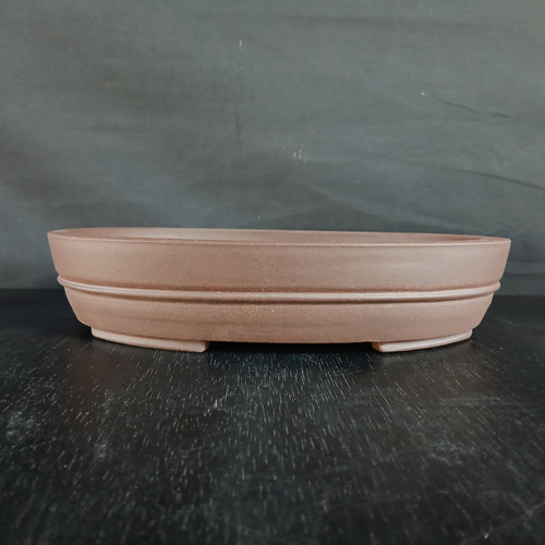 A detailed side-profile photograph of a 12-inch oval unglazed Yixing bonsai pot (No. 2286). The pot is crafted from high-quality, reddish-brown clay with a smooth, matte, and earthy finish characteristic of traditional Yixing pottery. Its shallow, oval-shaped body features a subtle horizontal decorative band or ridge that runs around the center of the exterior wall, adding a touch of classic elegance. The pot is supported by small, integrated feet at the bottom. It is displayed on a dark, slightly reflective surface against a dark, textured charcoal background, which highlights the pot's warm color and clean, symmetrical lines. This unglazed container is designed to provide a natural aesthetic for various bonsai tree species.