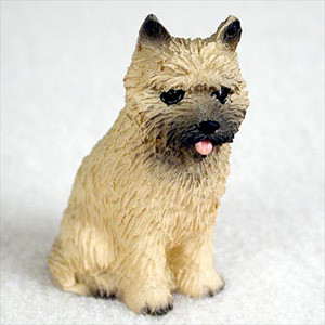 A detailed miniature bonsai tree figurine of a red Cairn Terrier sitting attentively. The figurine is crafted with a shaggy, textured coat in a light tan or "red" hue, mimicking the wire-haired appearance of the breed. It features a dark grey muzzle, a small black nose, and dark, watchful eyes. Its small, pointed ears are held upright with dark tips, and a tiny pink tongue peeks out playfully from its mouth. The dog is positioned in a seated pose on a soft, white, textured background, highlighting its small scale and fine details, making it a perfect accessory for a bonsai display or miniature garden.