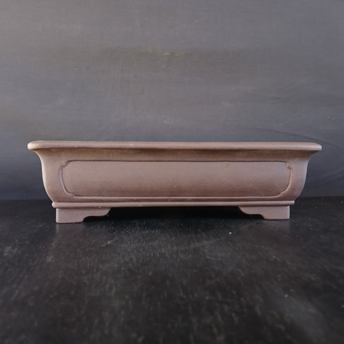 A detailed studio photograph of a 12-inch unglazed rectangular Yixing bonsai pot, model No. 2291-1 from Bonsai Outlet. The pot is crafted from high-quality, reddish-brown unglazed clay with a smooth, matte finish. It features a traditional design with a slightly flared top rim and a long, recessed rectangular panel on the front face for a subtle decorative touch. The vessel is supported by four elegantly carved, block-style feet that provide height and stability. It is captured from a straight-on perspective, sitting on a dark, textured wooden surface against a plain, dark gray background, which highlights the pot's clean lines, earthy tones, and professional craftsmanship.