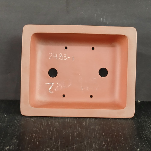 A top-down interior view of a 6-inch rectangular bonsai pot made from unglazed Yixing ceramic, showcasing its matte, reddish-brown clay finish. The pot features a wide, flat rim and an interior base designed for optimal plant health, containing two large circular drainage holes positioned centrally on either side. Four smaller holes for bonsai training wire are placed symmetrically near the corners of the base. Handwritten in white on the interior bottom is the model number "2483-1" along with other faint, indistinct markings. The pot is set against a dark gray background and rests on a dark, textured wood-like surface, emphasizing the earthy tones and functional design of traditional Yixing pottery.